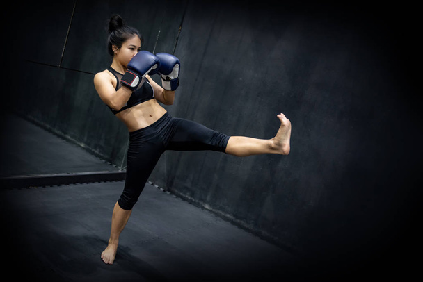 Jeune boxeuse asiatique avec des gants de boxe bleus posant garde et coups de pied dans le gymnase d'exercice. Cours d'arts martiaux ou de boxe féminine sur fond noir en studio. Concept de kickboxing
 - Photo, image