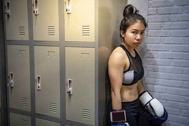 Mujer joven asiática boxeadora con guantes de boxeo de pie junto al armario en el vestuario. Concepto de clase de boxeo femenino
 - Foto, Imagen