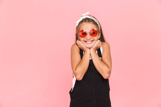 Cheerful little girl wearing dress and sunglasses standing isolated over pink background, looking at camera - Fotografie, Obrázek