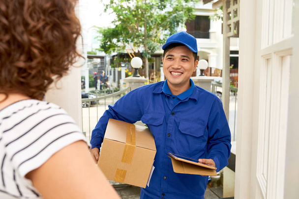 Smiling Asian courier with cardboard box talking to customer - Foto, afbeelding
