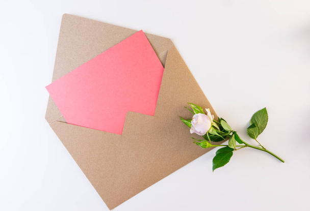 Λουλούδι της σειράς. Απαλό μοβ τριαντάφυλλο με πράσινα φύλλα σχετικά με craft φάκελο με κενό κομμάτι ροζ χαρτί σε άσπρο φόντο. Χώρο αντίγραφο για το κείμενο. - Φωτογραφία, εικόνα