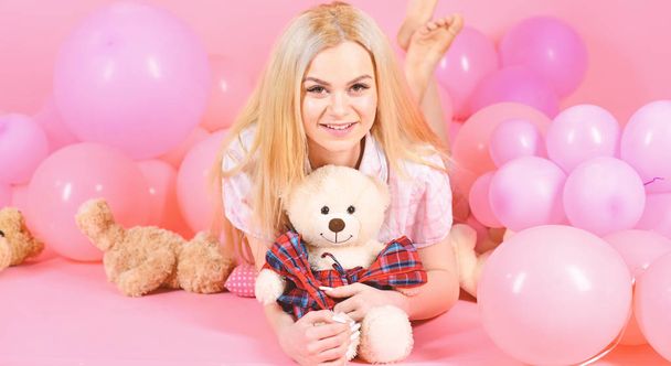 Blonde on smiling face relaxing with teddy bear toy. Woman cute celebrate birthday with balloons. Girl in pajama, domestic clothes lay near air balloons, pink background. Birthday girl concept - Zdjęcie, obraz