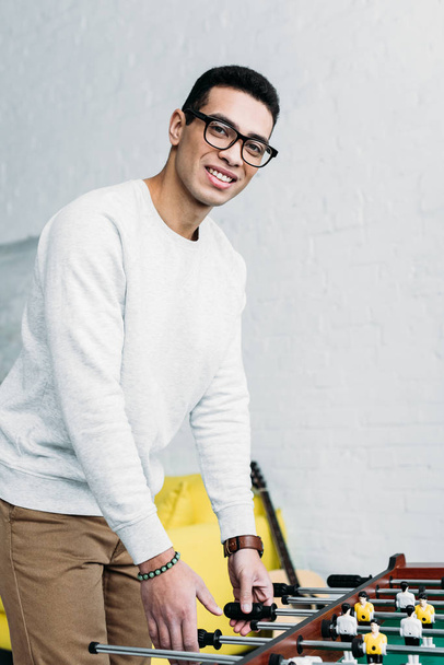 hymyilevä sekarotuinen nuori mies lasit pelaa pöytäjalkapalloa ja katsot kameraa
 - Valokuva, kuva