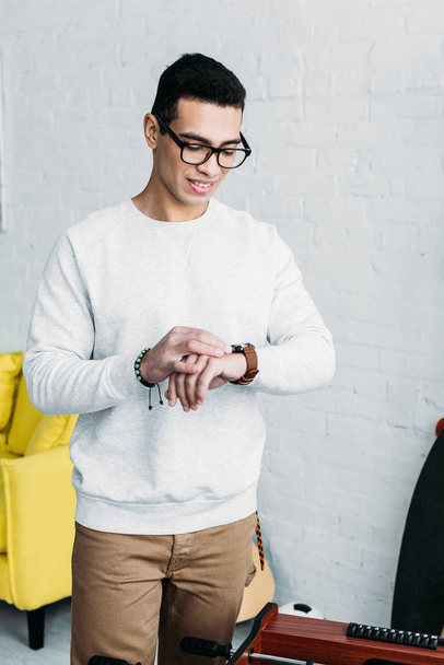 handsome mixed race man in white sweatshirt checking time on wristwatch - Фото, изображение