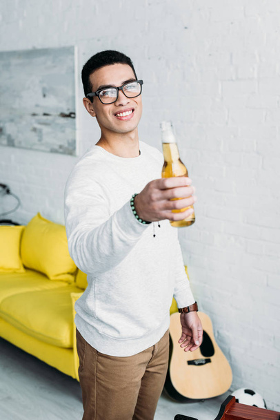 smiling handsome mixed race man holding bottle of beer and looking at camera - Foto, Bild