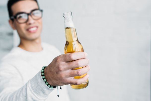 smiling mixed race man in glasses holding bottle of beer in stretched hand - Photo, image