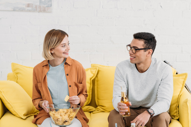 interracial couple talking and enjoying snacks and drinks while sitting on yellow sofa  - Photo, Image