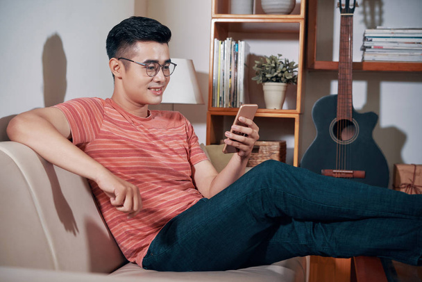 Веселий молодих в'єтнамський чоловік, сидячи на дивані у себе вдома і читання новин на смартфоні - Фото, зображення