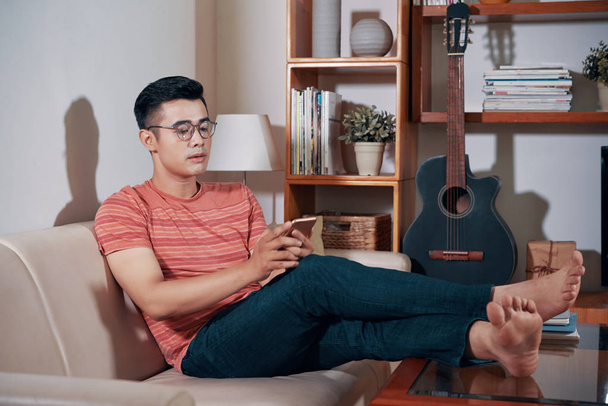 Handsome young Vietnamese man resting on sofa and reading text messages in his phone - Foto, imagen