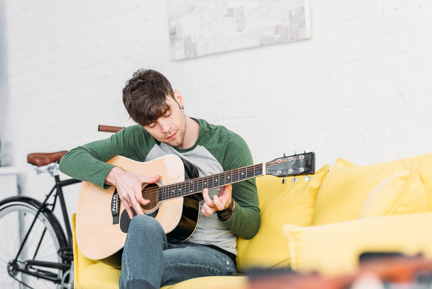 handsome young man playing acoustic guitar while sitting on yellow sofa - Foto, Imagen