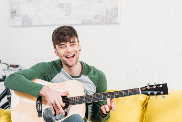joven tocando la guitarra acústica mientras está sentado en un sofá amarillo
 - Foto, Imagen