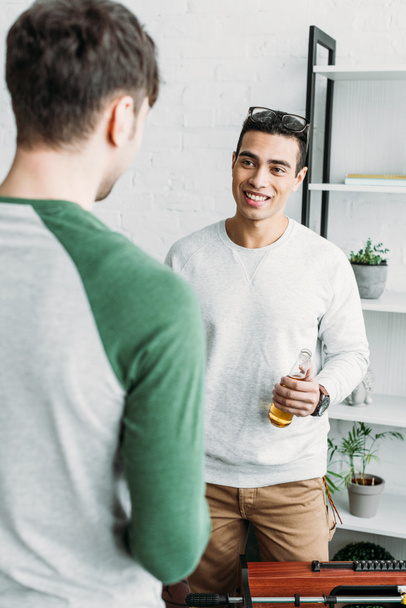 handsome mixed race man holding bottle of beer and talking with friend - Fotó, kép