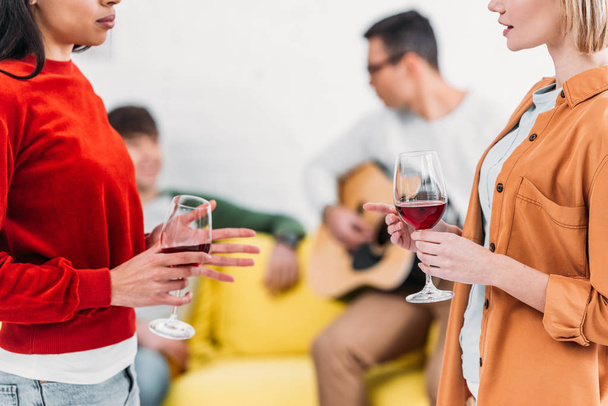 multicultural girls holding wine glasses and talking, men sitting on yellow sofa on background  - Фото, зображення