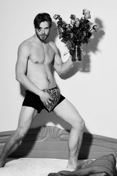 handsome bearded sexy macho man with stylish beard standing in underwear on bed near red blanket holding orange roses and decorative valentine heart with muscle torso on athletic body - Photo, image