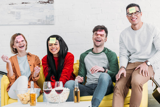 amigos multiculturais rindo com etiquetas apelido engraçado na testa sentado no sofá amarelo
 - Foto, Imagem