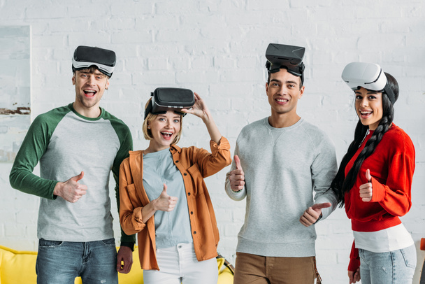 sonrientes amigos multiétnicos poniéndose auriculares de realidad virtual y mostrando los pulgares hacia arriba
 - Foto, Imagen
