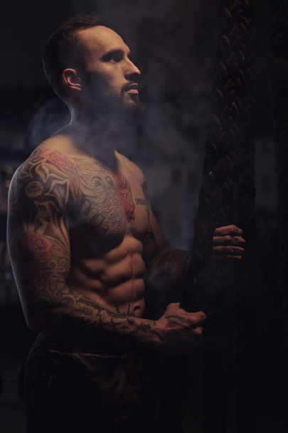 Shirtless, bearded, tattooed athlete holding ropes and looking up in the gym - Фото, изображение