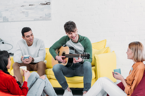 cheerful handsome man playing guitar to multicultural friends - 写真・画像