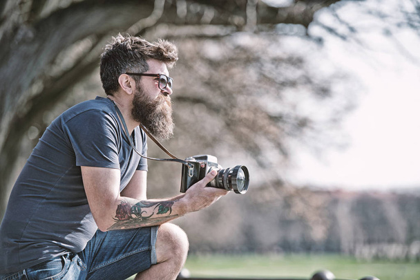 Bearded man works with vintage camera. Man with beard and mustache on concentrated face, branches on background, defocused. Man with long beard busy with shooting photos. Photographer concept - 写真・画像