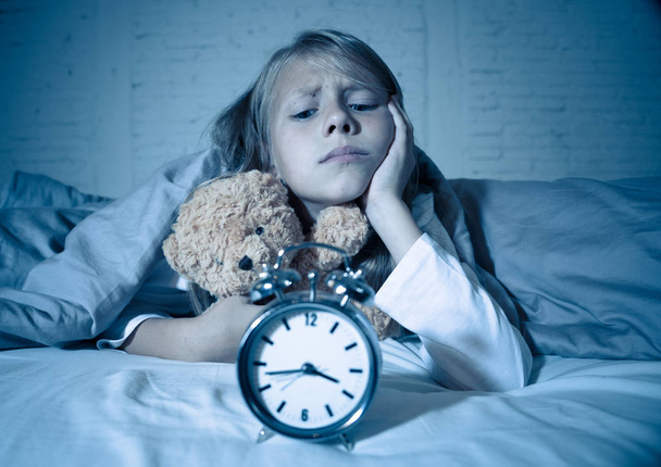 Cute sleepless little girl lying in bed showing alarm clock looking tired having sleeping troubles staying asleep at night or waking too early in Children Insomnia Anxiety and Sleep Disorders. - Foto, Bild