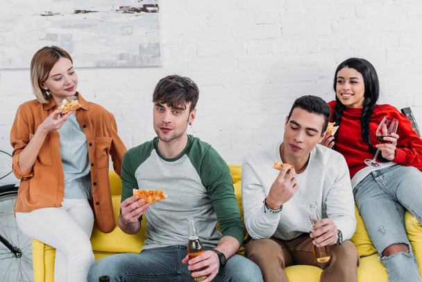 Happy πολυπολιτισμική φίλοι τρώγοντας πίτσα και απολαμβάνοντας ποτά ενώ κάθονται μαζί σε κίτρινο καναπέ - Φωτογραφία, εικόνα