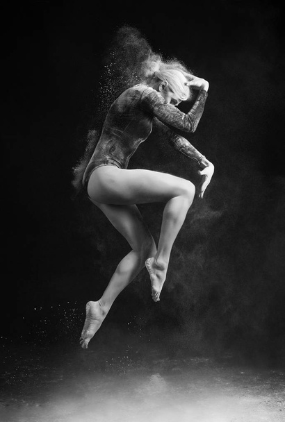 Beautiful slim girl wearing a gymnastic bodysuit covered with clouds of the flying white powder jumps dancing on a dark. Artistic conceptual and advertising black and white photo. Copy space - Photo, Image