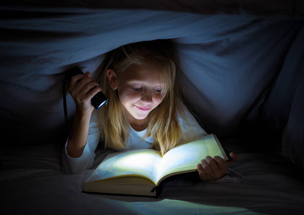 Happy smart girl reading a fairytale book lying in the dark under the cover bed holding a lantern in Reading skills Literature School Success and Education concept. - Foto, immagini