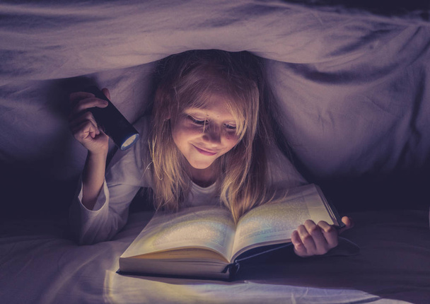 Gelukkig slim meisje lezen van een boek van de sprookjesachtige liggend in het donker onder het bed van de dekking met een lantaarn in lezing vaardigheden literatuur School succes en onderwijs concept. - Foto, afbeelding