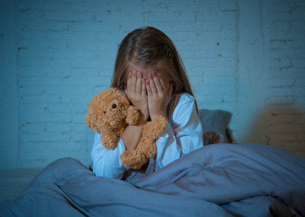 Scared little girl sitting in bed covering her face with hands holding her teddy in fear afraid of monsters in darkness in bedroom in Child nightmares imagination and psychological distress concept. - Valokuva, kuva