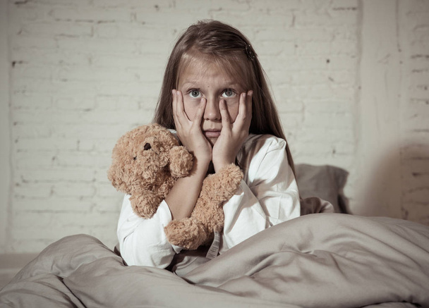 Scared little girl sitting in bed covering her face with hands holding her teddy in fear afraid of monsters in darkness in bedroom in Child nightmares imagination and psychological distress concept. - Fotografie, Obrázek