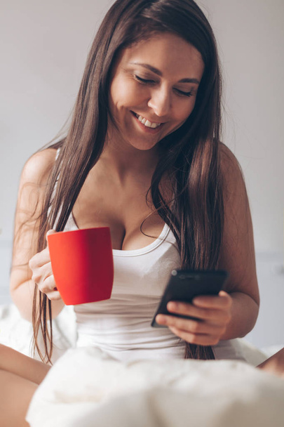 Young smiling woman drinking coffee at home in her bed and checking her smartphone. Red cup in hands - Valokuva, kuva