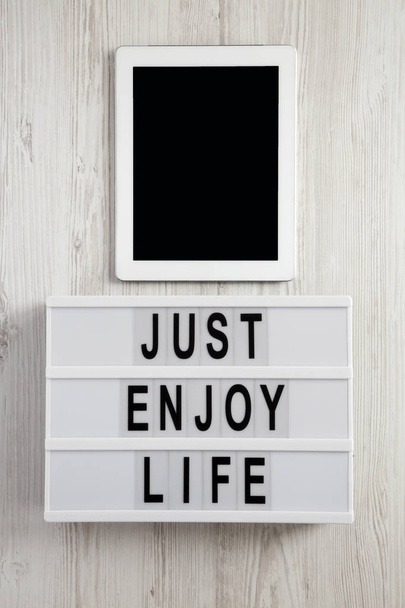 Tablero moderno con texto 'Just enjoy life', tableta con pantalla negra sobre una superficie de madera blanca, vista superior. Colocación plana, gastos generales
. - Foto, Imagen