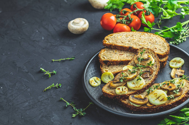 Delicious homemade whole grain rye sliced bread toast with fried cutting mushroom champignon and onion on black ceramic plate, herbs and vegetables, dark concrete table surface, copy space. - Photo, image