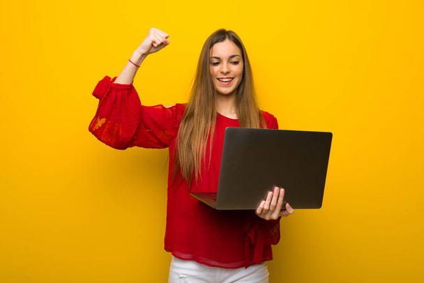 Jovem com vestido vermelho sobre a parede amarela com laptop e celebrando uma vitória
 - Foto, Imagem