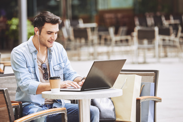 Jovem alegre sentado à mesa no café ao ar livre e trabalhando no laptop
 - Foto, Imagem
