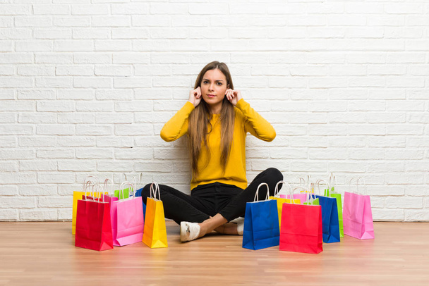 Chica joven con un montón de bolsas de compras que cubren las orejas con las manos. Expresión frustrada
 - Foto, imagen