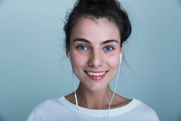 Close up of a beautiful young casual girl standing isolated over blue background, listening to music with earphones - Valokuva, kuva