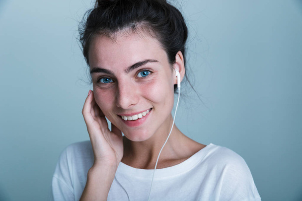 Close up of a beautiful young casual girl standing isolated over blue background, listening to music with earphones - Foto, imagen
