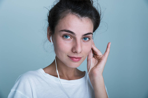 Close up of a beautiful young casual girl standing isolated over blue background, listening to music with earphones - Photo, image