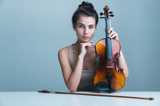Portrait d'une belle jeune femme assise à la table avec un violon isolé sur fond bleu
 - Photo, image