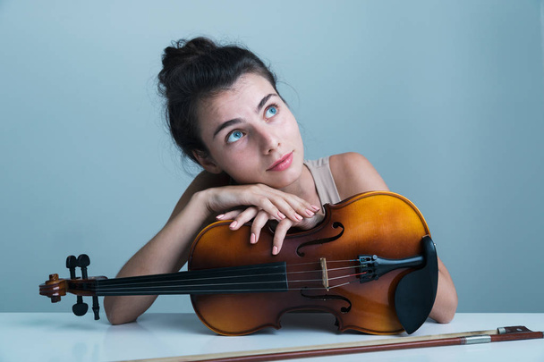 Portrait of a beautiful young woman sitting at the table with a violin isolated over blue background - Fotó, kép