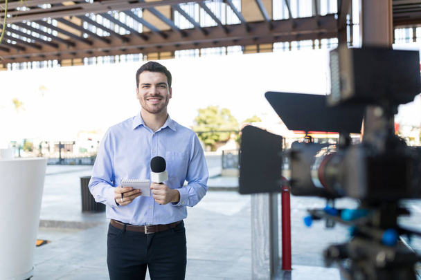 Porträt eines gut aussehenden lächelnden Nachrichtensprechers mit Mikrofon und Notizblock vor der Kamera - Foto, Bild