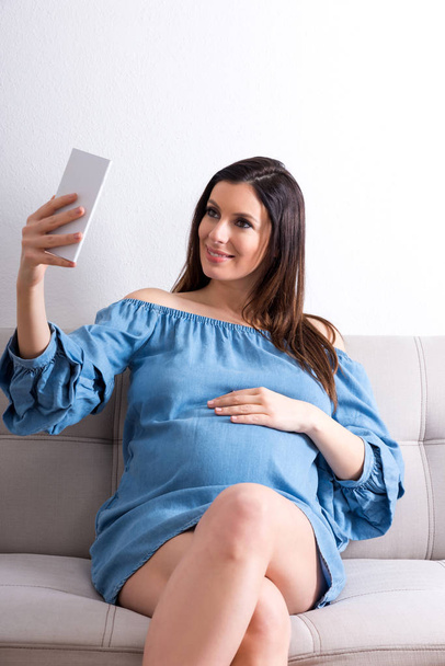 A pregnant woman sitting on a sofa and taking a selfie - Фото, зображення