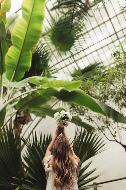 Joven hermosa mujer de vuelta en vestido blanco lanzando pequeño ramo de novia con grandes hojas verdes alrededor de pasar tiempo en acogedor invernadero
 - Foto, Imagen