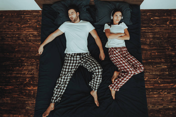 Couple is Lying on Bed. Couple is Young Beautiful Woman and Man. Man is Sleeping and Snores. Woman is Upset and Covering His Ears with Headphones. People in Home Interior. Top View. - Photo, Image