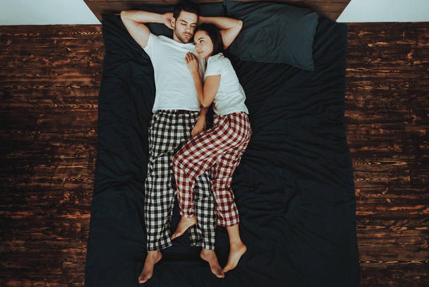 Couple is Lying on Bed. Couple is Young Beautiful Woman and Man. Woman is Hugging Man and Smiling. People is Wearing Pajama Pants and T-Shirts. Persons is Located in Home Interior. Top View. - Fotó, kép