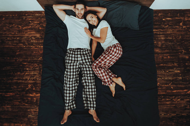Couple is Lying on Bed. Couple is Young Beautiful Woman and Man. Woman is Hugging Man. Persons is Looking Up and Smiling. People is Wearing Pajama Pants and T-Shirts. Home Interior. Top View. - 写真・画像