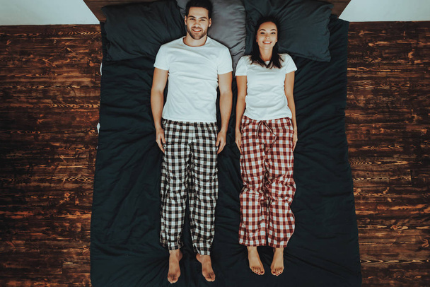 Couple is Lying on Bed. Couple is Young Beautiful Woman and Man. Persons is Looking Up and Smiling. People is Wearing Pajama Pants and T-Shirts. People is Located in Home Interior. Top View. - Photo, Image