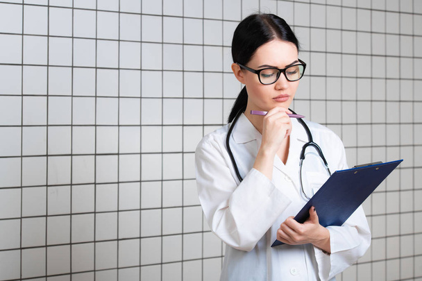 Jovem médica bonita em casaco cirúrgico branco com estetoscópio preto e suporte de papel azul em mãos em pé no consultório médico - Foto, Imagem