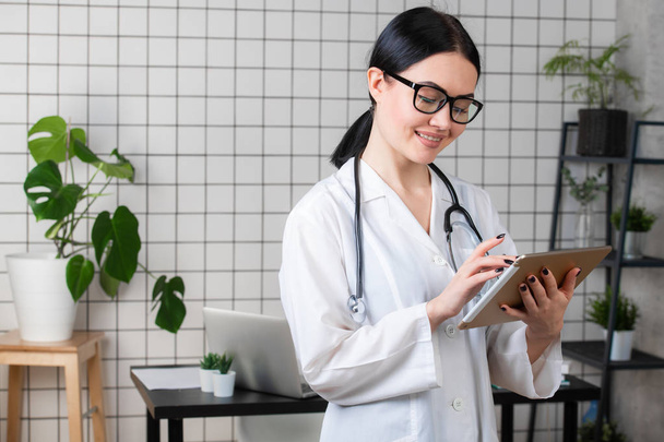 Női barna orvos tabletta, intelligens és kedves nő segítségével a szerkentyű és sztetoszkóp, a fehér office háttérben szemüveget visel - Fotó, kép